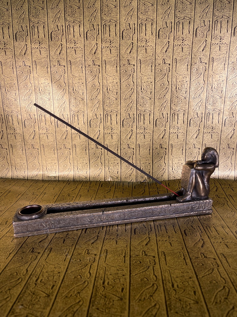 May include: A bronze incense burner with an Egyptian figure sitting on the side. The burner has a rectangular shape with a groove for incense sticks and a small round hole for ash. The figure is holding a hand to its chin and is looking down.