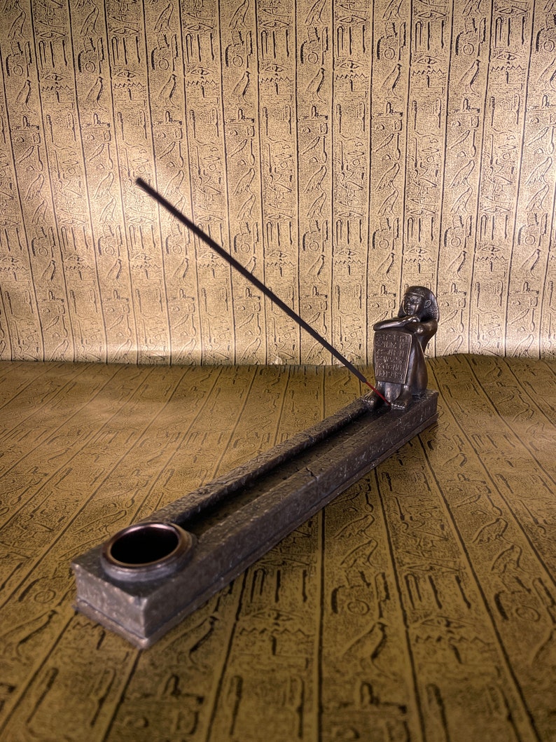 May include: A black incense stick burning in a gray stone incense holder shaped like an Egyptian pharaoh holding a tablet. The holder is on a gold patterned surface.