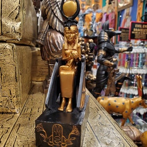 May include: Gold and black figurine of an Egyptian goddess with a sun disc headdress. The goddess is seated on a throne and holding a small figure in her lap. The figurine is on a black base with a gold symbol.