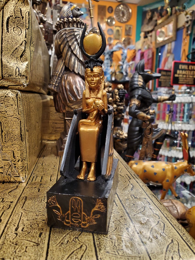 May include: Gold and black figurine of an Egyptian goddess with a sun disc headdress. The goddess is seated on a throne and holding a small figure in her lap. The figurine is on a black base with a gold symbol.