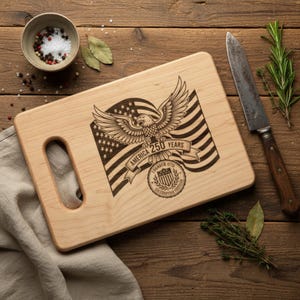 May include: A light-colored wooden cutting board with a handle, featuring an engraved design of an eagle, American flag, and text that reads "America 250 Years". A knife and herbs are placed nearby.