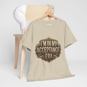 Könnte beinhalten: Beiges T-Shirt mit dem Text "I'M IN MY ACCEPTANCE ERA" in einem verzierten Design. Eine weiße Baseballkappe liegt auf einem Holzstuhl im Hintergrund.