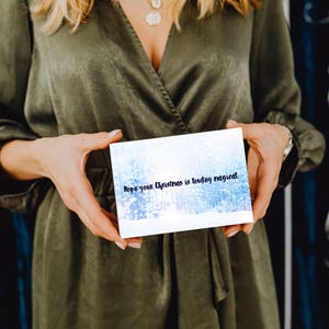 May include: A white rectangular card with a winter forest scene and the text "Hope your Christmas is lowkey magical" in blue script. The card is held by a person wearing a green dress.