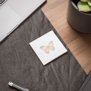Puede incluir: Un bloc de notas cuadrado blanco con un diseño de mariposa en acuarela. La mariposa tiene tonos amarillos, naranjas y verdes. Las notas están sobre una superficie gris oscuro, cerca de un portátil y una planta en maceta.