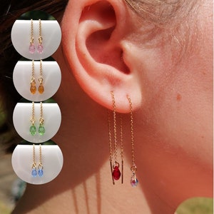 May include: Gold chain earrings with teardrop-shaped crystals in red and iridescent colours. Additional earrings in pink, orange, green, and blue are also shown. The earrings are displayed on a person's ear.
