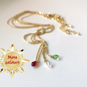 May include: A gold layered chain necklace with three small crystal charms: red, green, and clear. The necklace is on a white background with a gold sun-shaped sticker that says "More colours".