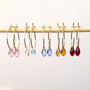 May include: A collection of gold hoop earrings, each adorned with a teardrop-shaped crystal. The crystals vary in color, including pink, clear, blue, amber, and red. The earrings are displayed on a wooden dowel against a white background.