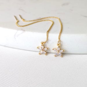 May include: A pair of gold dangle earrings with a small flower charm set with clear cubic zirconia stones.