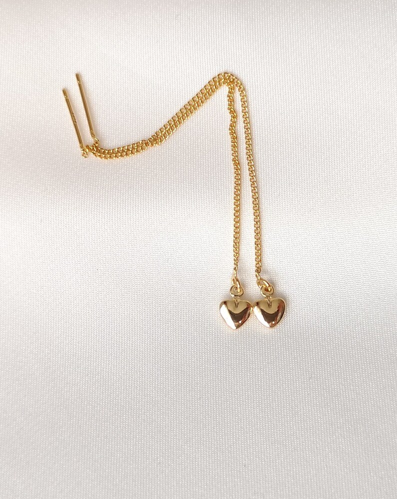 Pu&ograve; includere: Un paio di orecchini pendenti a forma di cuore dorati con una catena delicata.