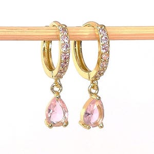 May include: Gold-coloured hoop earrings with pink gemstones. The hoops are encrusted with small pink stones, and each earring has a teardrop-shaped pink gemstone pendant. The earrings are displayed on a wooden dowel against a white background.
