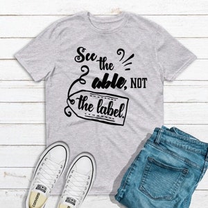 Może przedstawiać: Szary T-shirt z czarnym nadrukiem, na którym widnieje napis "See the able, not the label."