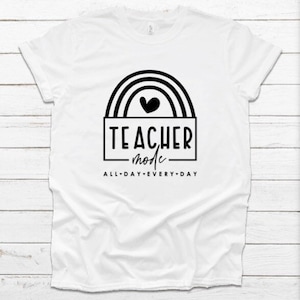Pode incluir: Camiseta branca com um design gráfico preto e branco. O gráfico apresenta um arco-íris com um coração no topo e o texto "TEACHER mode ALL-DAY EVERY-DAY".