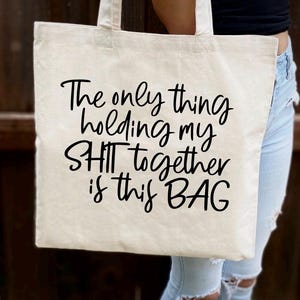 May include: A white canvas tote bag with black text that reads "The only thing holding my SH*T together is this BAG".