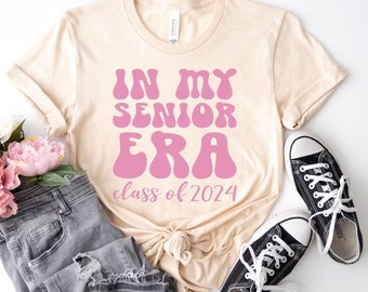 Puede incluir: Una camiseta beige con un gráfico rosa que dice "In My Senior Era Class of 2024".