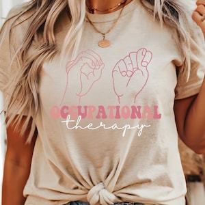 以下が含まれることがあります： 薄いベージュのTシャツに、アメリカ手話で「作業療法」をサインしている2つの手のピンク色のグラフィックが描かれています。「作業療法」という文字は、ピンク、赤、白のレトロなフォントで書かれています。