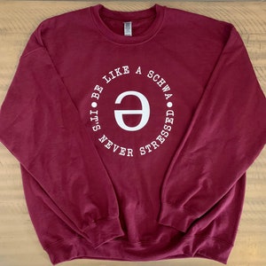 Könnte beinhalten: Burgunderfarbener Pullover mit einem weißen Kreis-Motiv, auf dem "It's never stressed. Be like a schwa." steht.