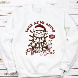 Puede incluir: Sudadera blanca con un gato gruñón con gorro de Papá Noel y luces navideñas. El texto dice "Look at me being all Festive & Shit" en una fuente festiva. Un diseño humorístico para las fiestas.