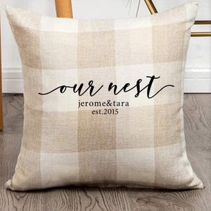 May include: A beige and white checkered throw pillow with the text "our nest jerome & tara est. 2015" in black.