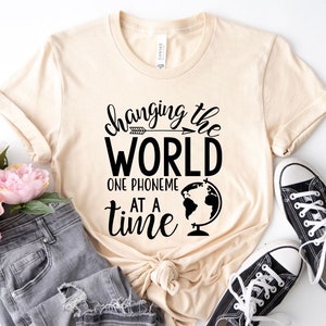 Op de afbeelding: Een beige T-shirt met zwarte tekst die "Changing the World One Phoneme at a Time" luidt. De tekst wordt vergezeld door een kleine pijl en een wereldkaart.