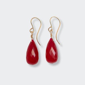 May include: A pair of teardrop-shaped earrings with faceted, deep red gemstones. The earrings have gold-colored, curved ear wires and are suspended from a small, wrapped wire detail. The gemstones have a polished, reflective surface.
