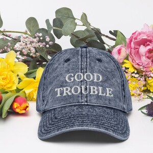 May include: A blue denim baseball cap with the text "GOOD TROUBLE" embroidered in white on the front.