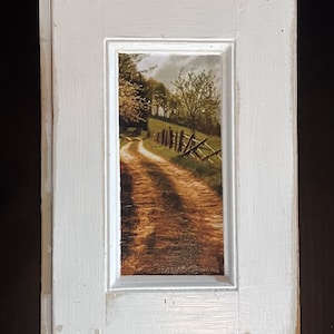 May include: A white wooden frame with a picture of a dirt road winding through a field with trees and a fence. The picture is in a rustic style.