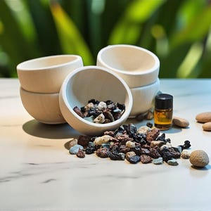 May include: A collection of small, light-colored wooden bowls, some filled with colorful stones. A small bottle of amber liquid and several smooth, rounded stones are also present. The items are arranged on a white surface with a blurred green background.