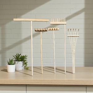 May include: A collection of small, wooden zen garden rakes in various designs. The rakes are light-colored, with different head shapes, including a straight edge, a toothed edge, and a fan shape. A small potted plant is visible in the background.