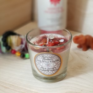 May include: A clear glass votive candle filled with white wax, topped with red jasper stones and a wick. The candle has a label that reads "Harmonious Amber Soy Wax Candle Red Jasper Problem Solver."