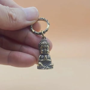 May include: A small, bronze-colored Buddha figurine keychain. The Buddha is in a seated meditation pose with hands clasped in prayer. The keychain ring is a decorative, open circle.