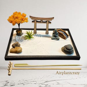 May include: A miniature Zen garden with a black frame, featuring a small tree with orange leaves, a wooden gate, a seated Buddha statue, and a small bridge. The sand is raked with circular patterns. Includes a rake and two tools.