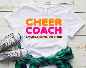 May include: White t-shirt with the text "CHEER COACH" in orange and pink letters. Below the text is the phrase "AMERICA RUNS ON SPIRIT" in brown letters.