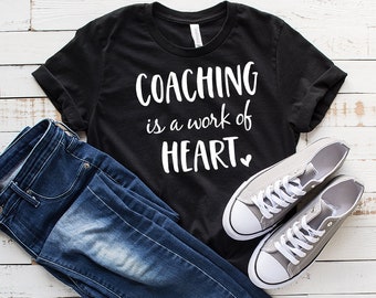 Peut inclure: T-shirt noir avec un texte blanc qui dit "COACHING is a work of HEART." Le t-shirt est posé sur une surface en bois blanc avec une paire de baskets grises et un jean bleu.