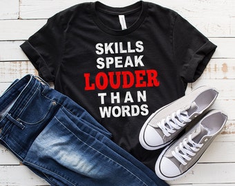 Peut inclure: T-shirt noir avec du texte blanc qui dit "Skills Speak Louder Than Words". Le texte "Louder" est en rouge. Le t-shirt est posé sur une surface en bois blanc avec un jean bleu et des baskets grises.