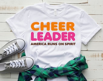 May include: White t-shirt with the text "CHEER LEADER" in orange and pink letters. The text "AMERICA RUNS ON SPIRIT" is printed in brown below the main text.