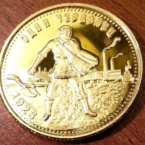 May include: A gold-colored coin with a depiction of a man holding a hammer and sickle in front of a factory on one side and a hammer and sickle with wheat stalks on the other side. The text on the coin reads "ОДИН УБРОНЬ" and "ПРОЛЕТАРИАТ ВСЕХ СТРАН, СОЕДИНЯЙТЕСЬ!" and "СССР 1923" and "СССР 1958".