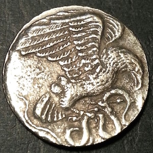 Puede incluir: Moneda de plata con una imagen detallada de un pájaro con las alas extendidas, posiblemente un águila, con una serpiente o serpiente debajo.