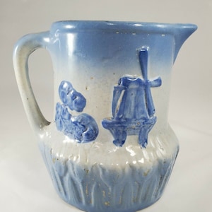 May include: Blue and white ceramic pitcher with a windmill and tulips design. The pitcher has a handle and a spout.