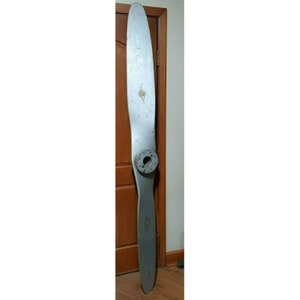 May include: A large, silver airplane propeller with a wooden hub. The propeller is leaning against a wooden door.