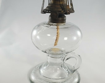Könnte beinhalten: Eine klare Glas-Öllampe mit einem Messingbrenner und einem Griff. Die Basis der Lampe hat ein Blumenmuster.