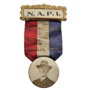 May include: A vintage commemorative ribbon with a gold-colored top piece that reads "N.A.P.I." The ribbon is red, white, and blue, with text indicating it was for a Washington D.C. committee in September 1915. A circular photo of a man in a hat is attached.