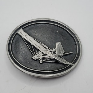 May include: A silver belt buckle with a black background and a detailed image of a small airplane.