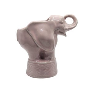 May include: A lavender ceramic elephant-shaped planter. The elephant has a trunk that forms a handle, and its head is tilted upwards. The planter has a decorative base with a geometric pattern.