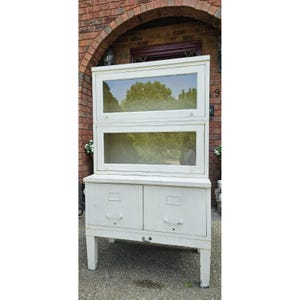 May include: A white vintage cabinet with glass-paned doors and drawers. The cabinet has a two-tiered display area above two drawers. The cabinet is supported by four legs. The cabinet is painted white and has a distressed finish.
