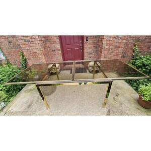 May include: A rectangular glass-top table with a gold-colored metal frame. The table is outdoors, with a brick wall and greenery in the background. The table's dimensions are approximately 72 inches long and 36 inches wide.