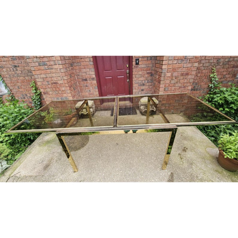 May include: A rectangular glass-top table with a gold-colored metal frame. The table is outdoors, with a brick wall and greenery in the background. The table's dimensions are approximately 72 inches long and 36 inches wide.