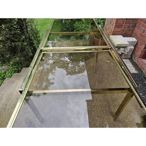 May include: A rectangular glass and brass table with a reflective surface. The table has a brass frame and legs. The glass top reflects the surrounding trees and sky. The table is on a concrete surface.