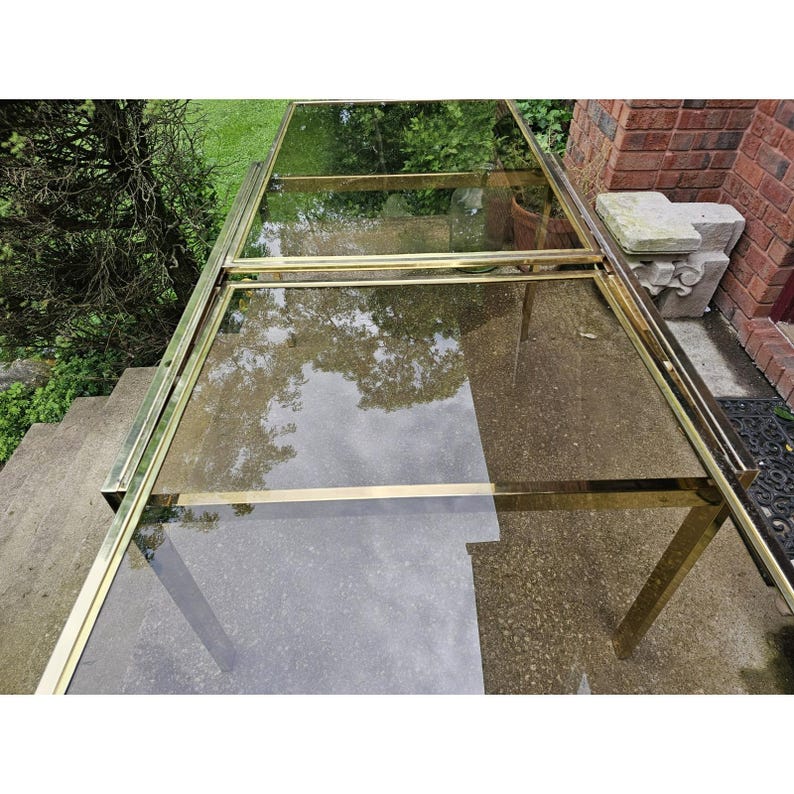 May include: A rectangular glass and brass table with a reflective surface. The table has a brass frame and legs. The glass top reflects the surrounding trees and sky. The table is on a concrete surface.