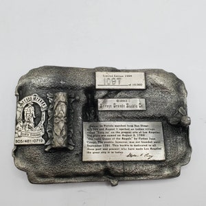 May include: A silver-toned metal belt buckle from Arroyo Grande Buckle Co. Features a rectangular shape with raised relief details and text. Includes the text "Limited Edition 1984" and "1097 of 10,000". The buckle has a historical theme.