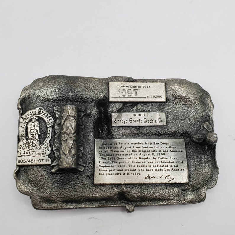 May include: A silver-toned metal belt buckle from Arroyo Grande Buckle Co. Features a rectangular shape with raised relief details and text. Includes the text "Limited Edition 1984" and "1097 of 10,000". The buckle has a historical theme.